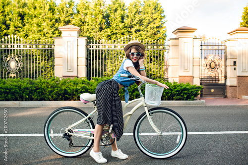 Attractive girl with long curly hair in hat posing with bike on road. She wears long skirt, jerkin, blue sunglasses. She is smiling to camera.