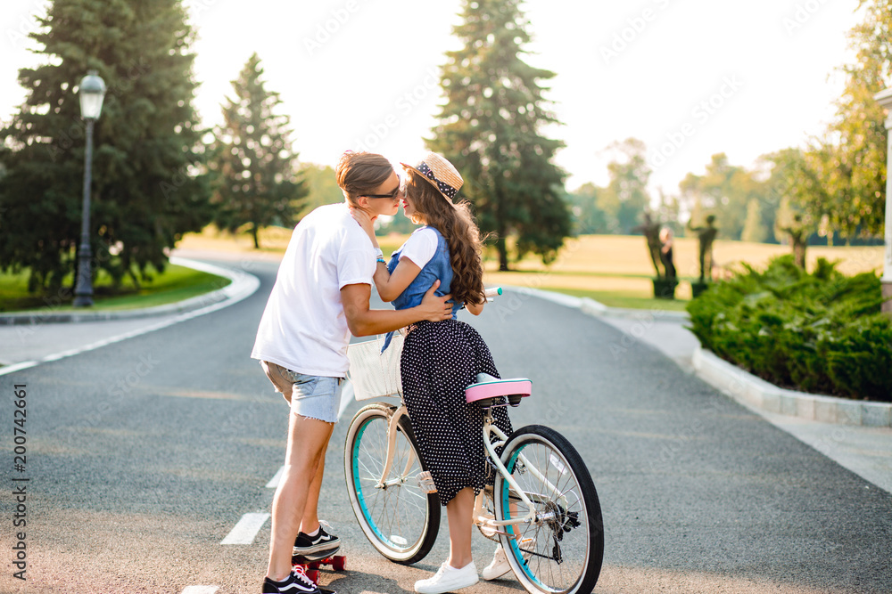Young couple in love on road on sunset. Pretty girl with long curly hair in hat and long skirt holds a bike, handsome guy on skateboard hugging and going to kiss her.