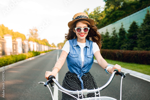 Cute girl with long curly hair in sunglasses driving a bike to camera on road. She wears long skirt, jerkin, hat. She looks happy.