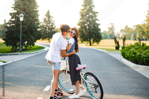 Wallpaper Mural Young couple of romantic people on road on sunset. Pretty girl with long curly hair in hat and long skirt holds a bike, handsome guy on skateboard hugging and looking at her. Torontodigital.ca