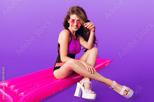 Attractive girl with long curly hair in pink sunglasses on purple background. She wears colorful swimsuit sitting on pink air mattress.