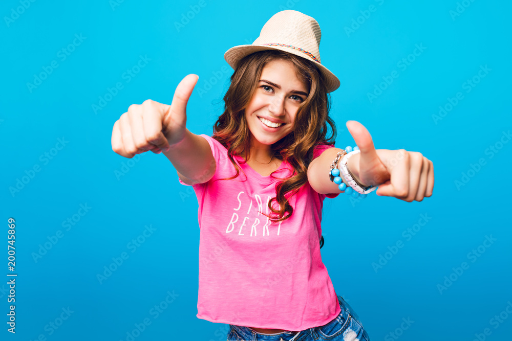 Pretty girl with long curly hair posing on blue background in studio. She wears pink T-shirt and hat. She shows hands to camera and looks happy. Pretty girl with long curly hair posing on blue background in studio. She wears pink T-shirt and hat. She shows hands to camera and looks happy.