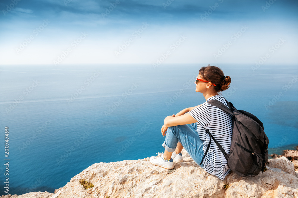 A girl traveler sits on a rock and admires the blue boundless sea, freedom, travel, unity with nature