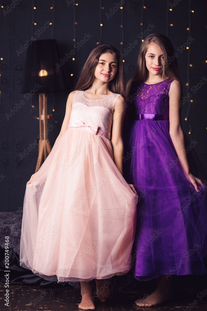 Cheerful girlfriends. Two long-haired girls in pink and purple dresses in studio on dark background with bokeh. Copy space.