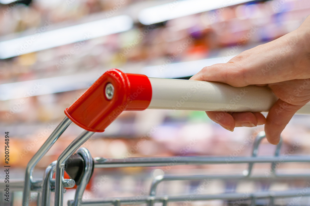 Close up view of a hand with pushcart in supermarket. The racks with the consumer goods in blur. Shallow depth of field