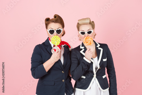 Cheerful girls couple with lollipops and glasses. Girls hide mouth by lollipops and eyes by glasses. Official women in non official style. Pinup styled women with curls in blue jackets and sunglasses photo