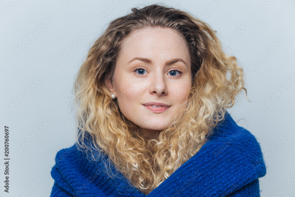 Portrait of pleasant looking curly woman with pleased delighted expression, has curly hair, wears casual warm sweater, looks directly into camera, isolated over light blue studio background. Portrait of pleasant looking curly woman with pleased delighted expression, has curly hair, wears casual warm sweater, looks directly into camera, isolated over light blue studio background.