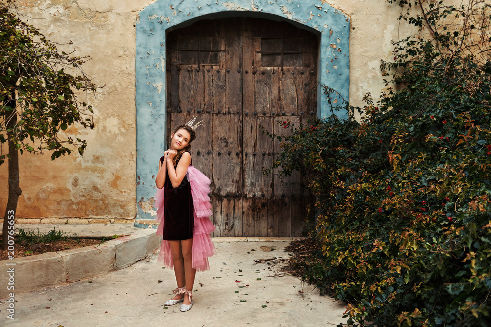 a sweet princess girl in a crown and a burgundy dress with a pink veil is pampered in front of an old house with an ancient wooden front door and a huge overgrown bush of dog rose