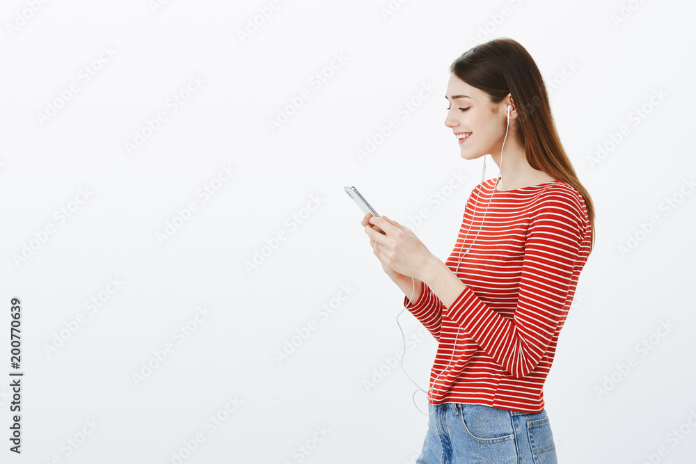 Girl recalling good memories, looking at photos in smartphone album. Portrait of pretty european female student, standing in profile, listening music in earphones or watching video in smartphone Girl recalling good memories, looking at photos in smartphone album. Portrait of pretty european female student, standing in profile, listening music in earphones or watching video in smartphone