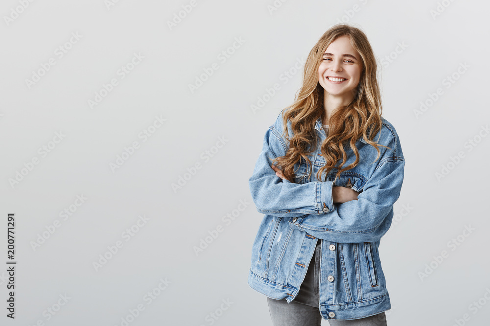 Happy to have great friends near. Portrait of grinning attractive caucasian female student with blond hair, wearing stylish denim jacket and standing with crossed hands, laughing over joke Happy to have great friends near. Portrait of grinning attractive caucasian female student with blond hair, wearing stylish denim jacket and standing with crossed hands, laughing over joke