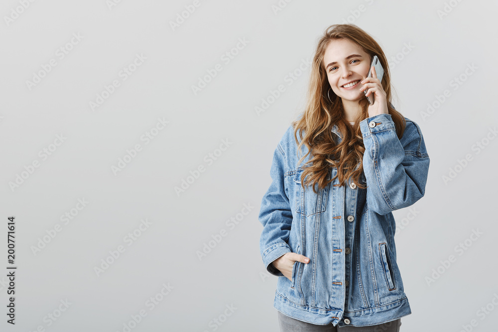 Girl is telling where is she standing. Portrait of carefree feminine caucasian blonde in denim jacket, holding hand in pocket, talking on smartphone with mom, standing over gray background pleased Girl is telling where is she standing. Portrait of carefree feminine caucasian blonde in denim jacket, holding hand in pocket, talking on smartphone with mom, standing over gray background pleased