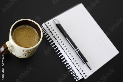 Coffee, notebook and pen on black wooden table. Coffee is an important drink in the workplace.