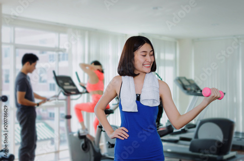 young asian pretty woman slim body doing exercises with dumbbell at biceps for good healthy in fitness gym at morning with people background, bodybuilder, lifestyle and sport exercise concept