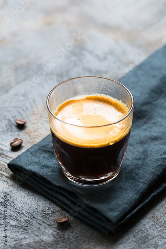 Glass cup of fresh coffee with thick golden foam froth on dark rustic background, espresso americano on black tissue with spoon of brown sugar and coffee beans, vertical copy space
