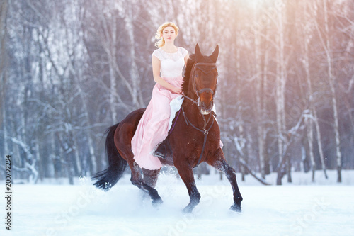 Young woman in rose dress riding her bay horse on winter field. Romantic or historical equestrian background with copy space