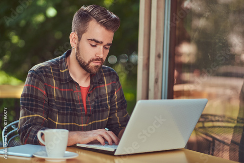 A handsome fashionable male freelancer with stylish haircut and beard, wearing fleece shirt, working on a laptop computer inside a cafe.