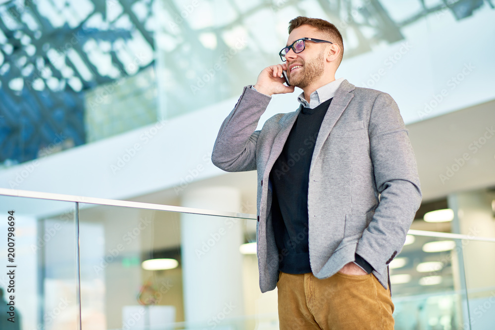 Smiling bearded manager wearing stylish clothes discussing project details with colleague on smartphone while standing at modern office lobby, portrait shot Smiling bearded manager wearing stylish clothes discussing project details with colleague on smartphone while standing at modern office lobby, portrait shot