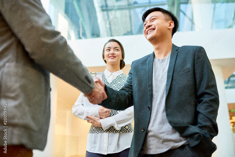 Low angle view of smiling Asian entrepreneur greeting his business partner with firm handshake while standing at spacious office lobby, pretty assistant manager seen behind him Low angle view of smiling Asian entrepreneur greeting his business partner with firm handshake while standing at spacious office lobby, pretty assistant manager seen behind him