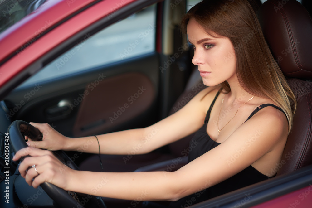 Concept: strong independent young woman in car. Beautiful serious girl sit behind the wheel at twilight. Outside look.