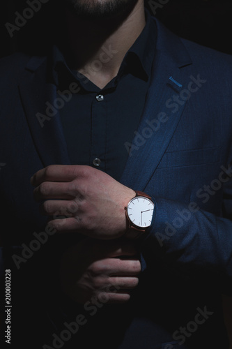 A dark portrait of a man dressed in an expensive suit, adjusting an elegant wrist watch