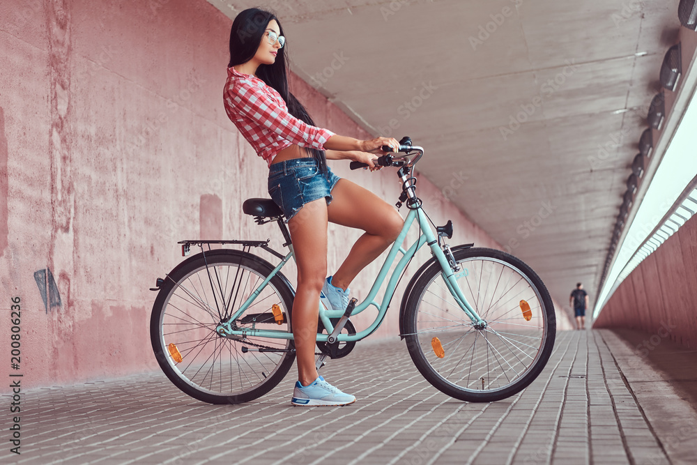 Naklejka premium Sexy stylish brunette girl wearing a pink flannel shirt and denim shorts in glasses, posing with city bike against a pink wall.