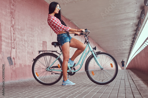 Sexy stylish brunette girl wearing a pink flannel shirt and denim shorts in glasses, posing with city bike against a pink wall.