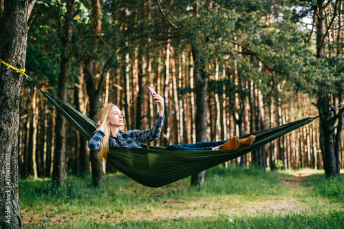Young beautiful blonde girl in hammock and making selfie. Pretty woman leisure lifestyle portrait at nature ountdoor. Female relax in forest. Adorable teen face expression. Tourism in summer camp.