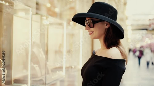 A beautiful stylish lady in a hat and sunglasses approaches the shop window of a boutique and looks at the goods with interest. Luxury jewelry store