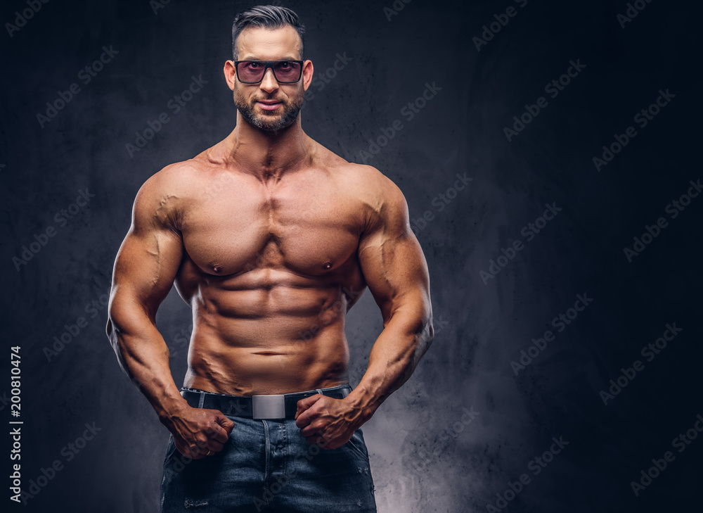 Portrait of a shirtless tall huge male with a muscular body with a stylish haircut and beard, in a sunglasses and jeans, posing in a studio.