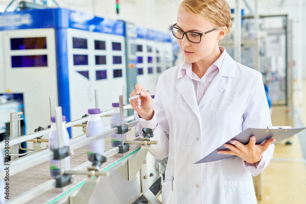 Portrait shot of pretty young worker wearing white coat standing at conveyor belt and controlling production process at modern soy milk factory, she holding pen and clipboard in hands Portrait shot of pretty young worker wearing white coat standing at conveyor belt and controlling production process at modern soy milk factory, she holding pen and clipboard in hands