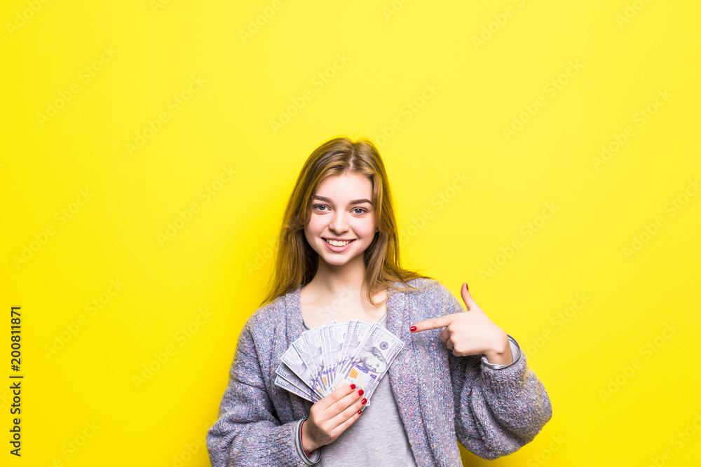 Joyful teenage girl with dollars in her hands pointed on them isolated on yellow background Joyful teenage girl with dollars in her hands pointed on them isolated on yellow background