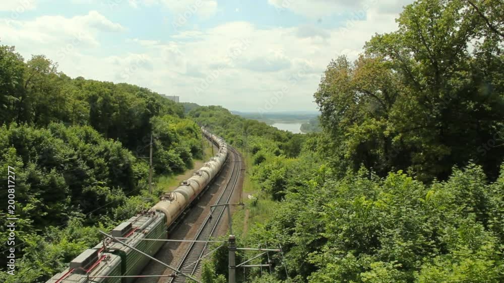 An electric locomotive,freight train carries oil tanks by two-sided railway along the river and green forest at hot summer day- top view. An electric locomotive,freight train carries oil tanks by two-sided railway along the river and green forest at hot summer day- top view.