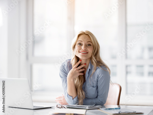 What can I help you. Portrait of polite young businesswoman is sitting at table in office. She is looking forward with joy and smiling