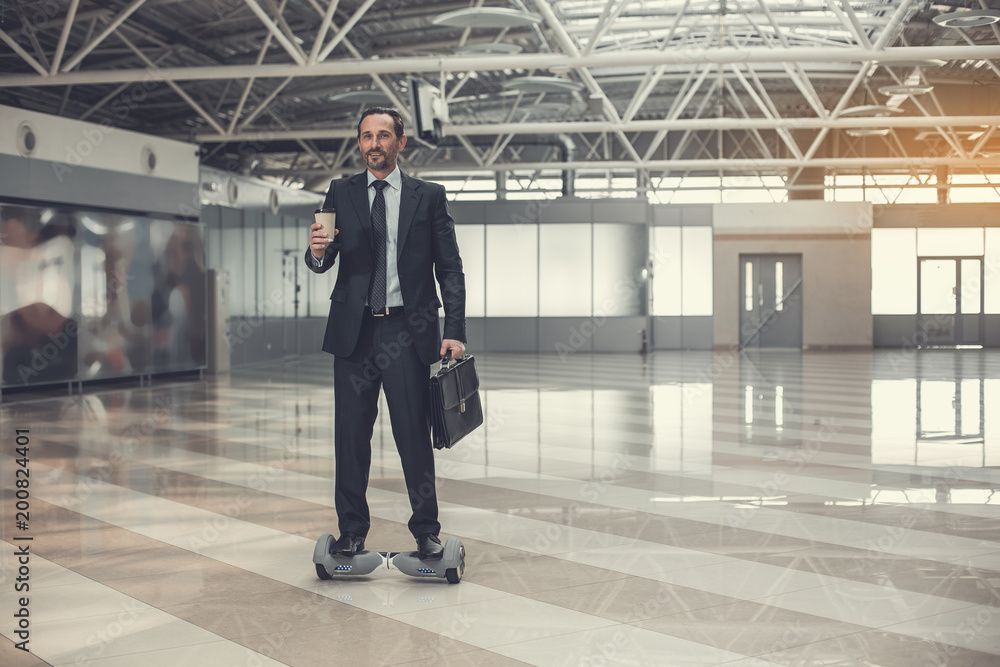 Full length portrait of satisfied employer tasting mug of hot liquid while going on gyroboard indoor. Cheerful male using gadget during break concept Full length portrait of satisfied employer tasting mug of hot liquid while going on gyroboard indoor. Cheerful male using gadget during break concept