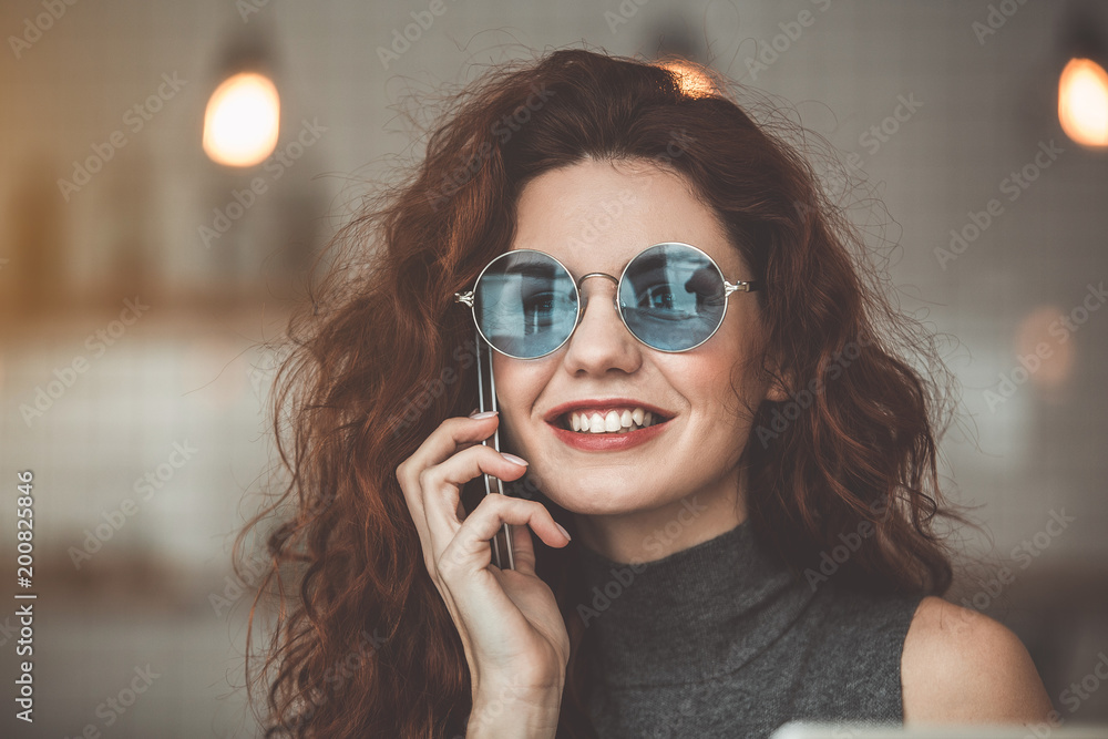 Portrait of excited young woman calling on mobile phone and smiling. She is wearing stylish sunglasses Portrait of excited young woman calling on mobile phone and smiling. She is wearing stylish sunglasses
