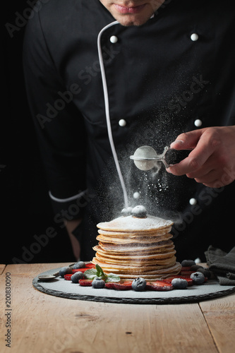 Serving pancakes with powdered sugar and berries. Chef man hand. Beautiful food still life. slightly toned image, dark black background photo