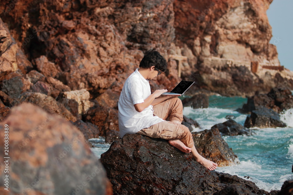 Young Asian man traveler using laptop on the rock of natural seashore. Vacations summer concept. Young Asian man traveler using laptop on the rock of natural seashore. Vacations summer concept.