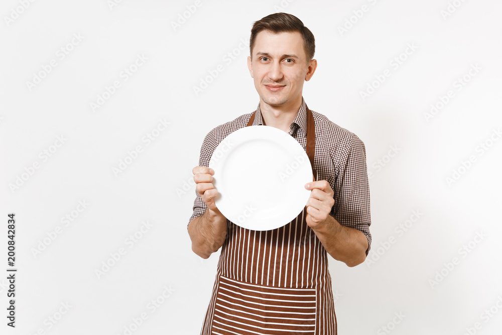 Young man chef or waiter in striped brown apron, shirt holding white round empty clear plate isolated on white background. Male housekeeper or houseworker. Domestic worker copy space for advertisement