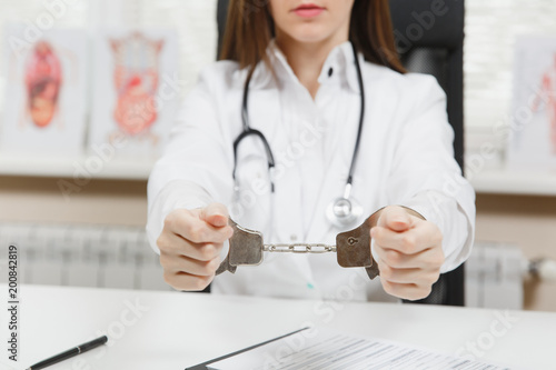Close up arrested female doctor sitting at desk with medical documents in light office in hospital. Woman in medical gown  stethoscope  hands with handcuffs in consulting room. Medicine  law concept.