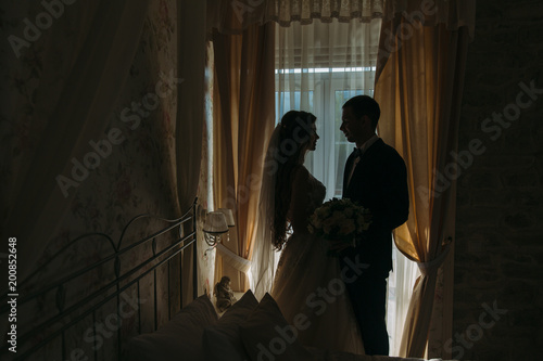 Newlyweds at wedding day. happy luxury bride and groom standing at window light in rich room, tender moment.