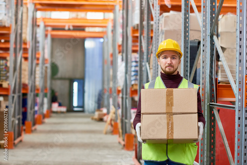 Portrait of young warehouse worker holding cardboard box and looking at camera while doing shipping and deliveries, copy space
