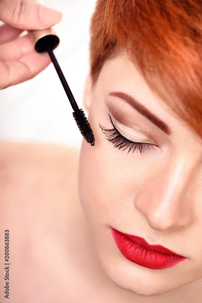 Makeup artist applies mascara brush. Beautiful young woman with short red hair. Makeup detail.