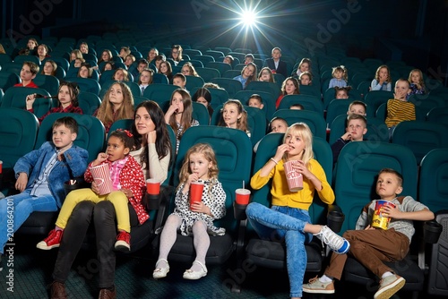Photo of emotional people watching film in the new cinema hall. There are adults and children sitting on comfortable sits wearing colorful clothes, keeping fizzy drinks, eating popcorn and smiling.