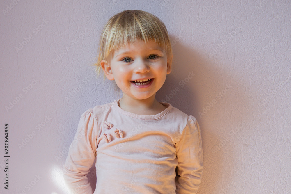 Happy little blond girl stands leaning against the wall in a sunny room at home. She looks at the viewer and laughs joyfully. Close up shot. Copy space. Happy little blond girl stands leaning against the wall in a sunny room at home. She looks at the viewer and laughs joyfully. Close up shot. Copy space.
