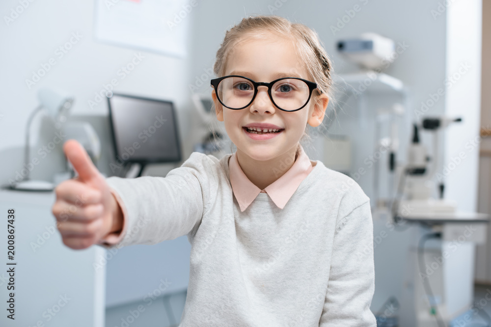smiling little child in eyeglasses showing thumb up in optical clinic smiling little child in eyeglasses showing thumb up in optical clinic