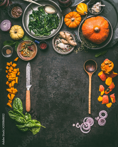 Autumn seasonal food, eating and cooking background with pumpkin.  Dark rustic kitchen table  with tools, bowls, spoons, knife, whole pumpkin and vegetables, top view, frame. Vegetarian food