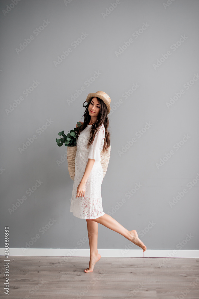 beautiful young brunette girl in white short dress, wearing hat, with summer bag in hand on gray background. Let's go shopping beautiful young brunette girl in white short dress, wearing hat, with summer bag in hand on gray background. Let's go shopping