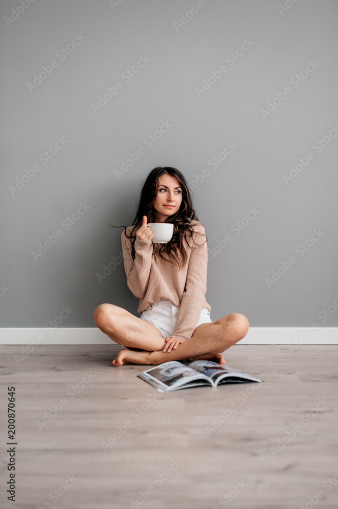 beautiful young brunette girl in white short shorts sitting on floor on gray background and drinking coffee beautiful young brunette girl in white short shorts sitting on floor on gray background and drinking coffee