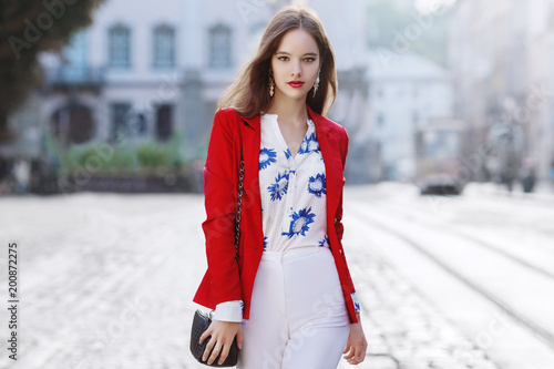 Outdoor portrait of a young beautiful confident woman posing in the street of european city. Model wearing stylish white blouse with flower print, high waist pants, red jacket. Female fashion concept