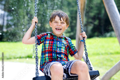 Funny kid boy having fun with chain swing on outdoor playground while being wet splashed with water
